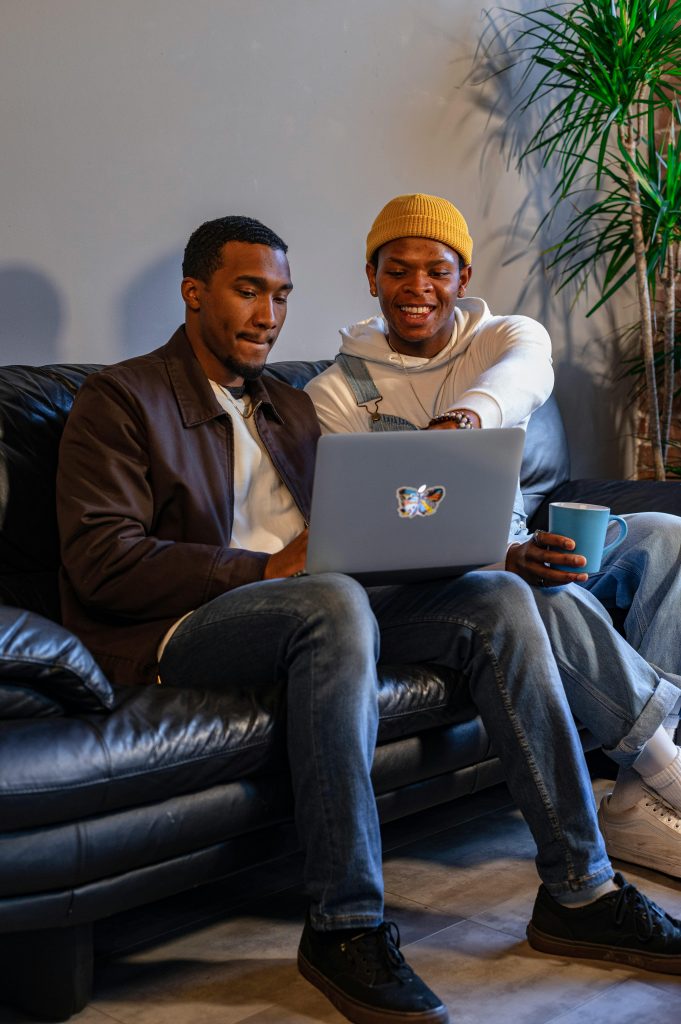 Two friends sitting on a sofa enjoy a moment of laughter while looking at a laptop indoors.