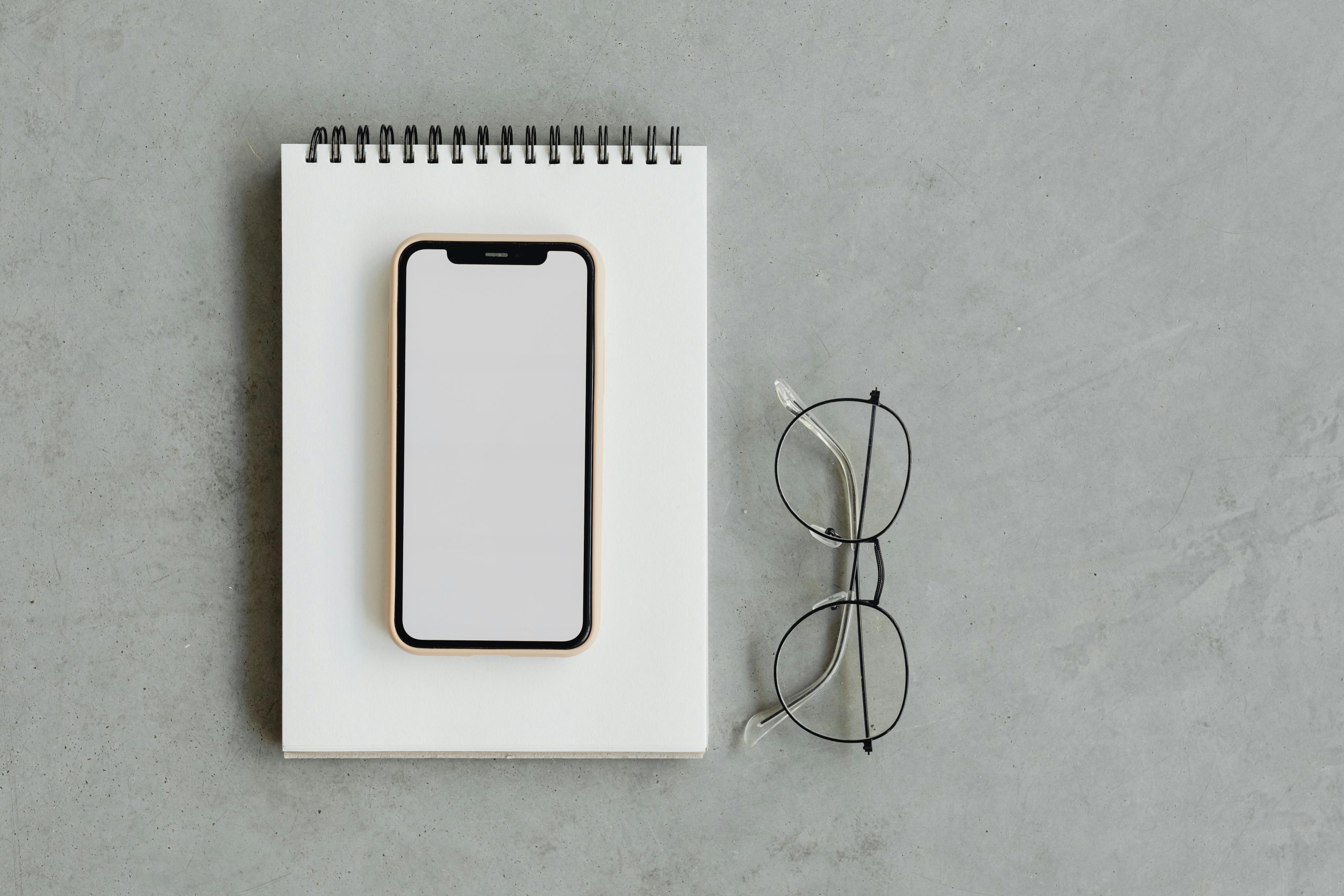 A minimalist flat lay composition featuring a smartphone, notepad, and eyeglasses on a gray surface.