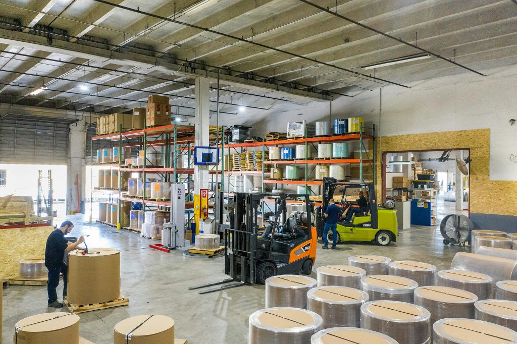 Interior view of a busy warehouse with forklifts and workers handling large rolls of material.